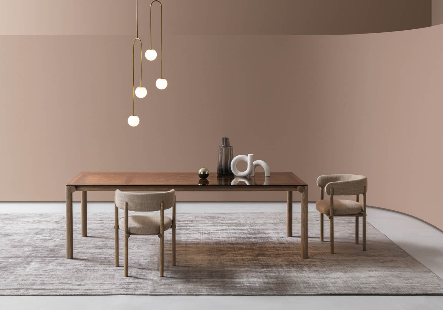 Tavolo in legno con piano in vetro bronzato, due sedie imbottite beige, lampada a sfere dorata e tappeto grigio su parete rosa